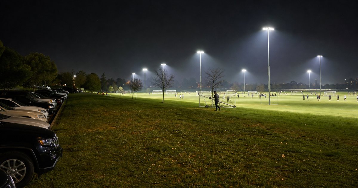 A public park with three adjacent lit soccer fields at night, teams warming up on each field, a groundskeeper moving portable goals, parked cars along the edge of the lot