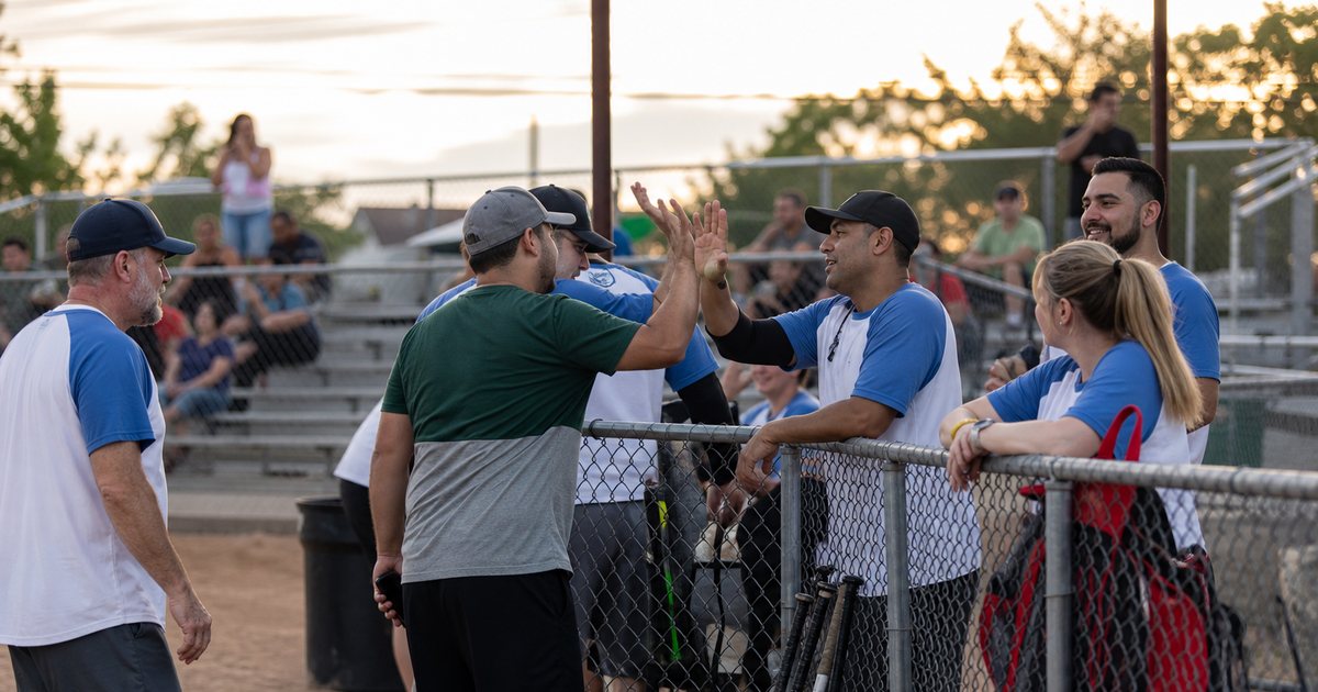 Adult recreational softball players gathered around a dugout fence at a public park field in the evening, gloves and bats leaning against the fence, some players high-fiving, bleachers in the background