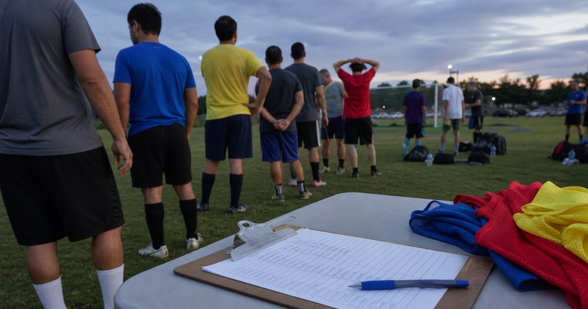 A clipboard with a handwritten sign-up sheet on a folding table beside a grass soccer field at dusk, adult players in mismatched jerseys lining up, a pen and a stack of colored pinnies nearby